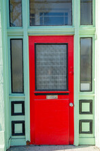 The Exterior Facade Of A Vintage Shop With An Orange Wooden Door With A Glass Window, Mailslot And Door Handle. The Door Is On A Lime Green Building With Black Trim. There's A Transom Window Over Door