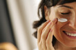 © Look! - Close-up happy young caucasian woman smiling with her eyes closed applying cream on her face. Brunette moisturizes her skin with cosmetics. Home spa care concept