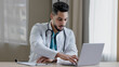 © Yuliia - Concentrated arabian man doctor practitioner sit at clinic cabinet use computer chatting distant in medical social app give online consultation writing recipe prescription diagnose search information