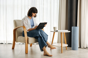 Positive young caucasian brunette woman working on tablet sitting indoors in morning. Girl wears shirt and jeans, reads messages on device. Technology concept