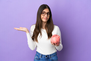 Wall Mural - Young caucasian woman holding a piggybank isolated on purple background having doubts while raising hands