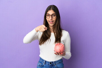 Wall Mural - Young caucasian woman holding a piggybank isolated on purple background surprised and pointing front