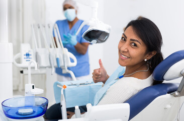 Happy latin american woman sitting in dental chair after teeth cure in modern clinic, gesturing thumbs up