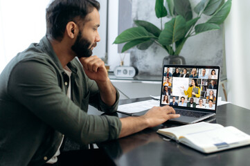 Wall Mural - E-learning, Online education, webinar. Focused successful smart mixed race man listening to an online lecture, taking notes in notebook, on a laptop screen, a teacher and a group of multiracial people