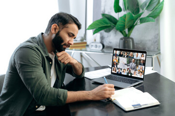 Wall Mural - Financial educational course. Focused successful smart mixed race man listening to an online financial lecture, taking notes in notebook, on a laptop screen,a teacher and a group of multiracial people