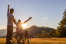 Silhouette Of Joyful Couple In Wheelchair Raised Hands At Sunset