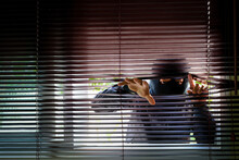 Unrecognizable Man Wearing Balaclava Face Mask Looking Through Venetian Blind. Keep Your Home Secure During Your Summer Holiday Concept.