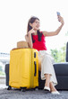 © Bangkok Click Studio - Asian young happy cheerful female traveler in casual vacation outfit with sunglasses sitting smiling on cozy sofa in living room holding smartphone taking selfie photo with yellow trolley luggage