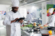Head chef with modern touchscreen tablet device searching for gourmet cuisine dish recipe while colleagues chopping fresh vegetables. Gastronomy expert with handheld device standing in kitchen.