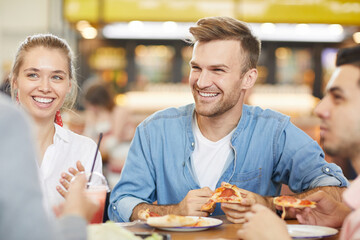 Wall Mural - Group of cheerful excited friends sitting at table in fast food restaurant and eating pizza while chatting