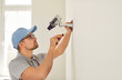 Professional male surveillance camera installer mounts it on wall inside new home or office. Side view of serious young man in t-shirt, cap and in glasses with screwdriver fastens cctv camera.
