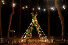 Festive String Lights Illumination On Boho Tipi Arch Decor On Outdoor Wedding Ceremony Venue In Pine Forest At Night. Vintage String Lights Bulb Garlands Shining Above Chairs At Summer Rural Wedding.