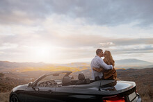 Young Couple In Love Kissing Near Convertible Car At Roadside With Romantic Sunset. Rear Concept