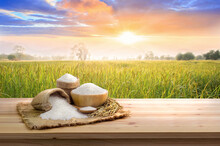 Asian Uncooked White Rice With The Sunset Rice Field Background And Burlap Sack On Wooden Table. Rice Grains Healthy Food