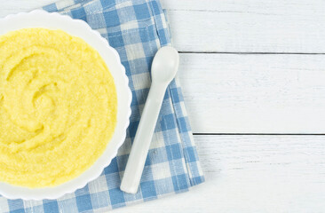 Wall Mural - Corn porridge for a baby made of ground cereals in a white bowl on a blue cloth napkin on a white wooden background. Space for the text. The first complementary food of a child, baby nutrition.