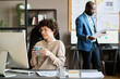 © pressmaster - Young businesswoman drinking tea and looking at computer monitor while sitting at her office desk with man working in background