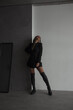 © NASTYA PALEHINA - teen fashion leisure. young girl in black dress-jacket and over the knee boots stands casual and looks apart with fun smile near white and gray studio wall background. lifestyle concept, free space