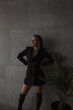 © NASTYA PALEHINA - teen fashion. young girl in black slim dress-jacket and over the knee boots stands fashion with hands on waist and looking apart on gray loft wall background with plant . lifestyle concept, free space