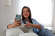 © Evrymmnt - Portrait of young brunette woman wearing mom jeans sitting on the couch texting and smiling. Joyful female having fun video phone call at home. Background, copy space, close up.