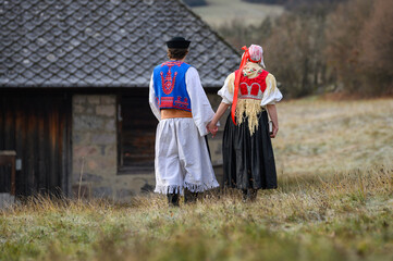 Sticker - A couple dressed in traditional folk costume. Slovak costume in autumn nature. Old country cottage in the background. Young couple in folk costume walking in the garden