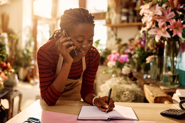 Wall Mural - African American florist writing order while talking to customer over the phone at flower shop.