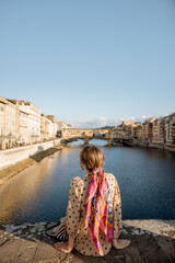 Wall Mural - Young woman enjoys beautiful view on famous Old bridge in Florence, sitting back on the riverside at sunset. Female traveler visiting italian landmarks. Stylish woman wearing dress and colorful shawl