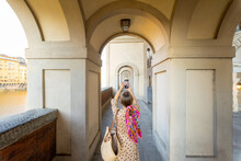 Young Woman Photographing On Phone Beautiful Archway, Visiting Famous Italian City Florence. Female Tourist Enjoys Italian Old Architecture. Woman Dressed In Italian Style With Colorful Scarf In Hair