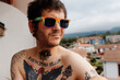 © Alberto - portrait of a caucasian man, with a beard and tattooed, wearing gay pride rainbow sunglasses on the terrace of his home. Real people, diverse and alternative.