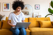 © Daniel - Young African american woman sitting on the couch using laptop working at home. Copy space.