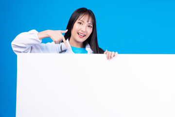 Wall Mural - Positive cheerful asian woman showing white placard empty copy space standing over isolated blue background. Teenager girl pointing to blank Banner, advertisement, sign board presentation concept.