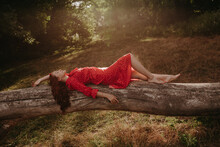 A Young And Beautiful Woman In A Red Summer Dress With White Flecks Lying On A Fallen Dry Tree Trunk In The Middle Of The Forest