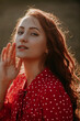 Portait of beauty young redhead woman with long curly thick hair dressed in red shirt dress with white dots posing on a camera in sunlight