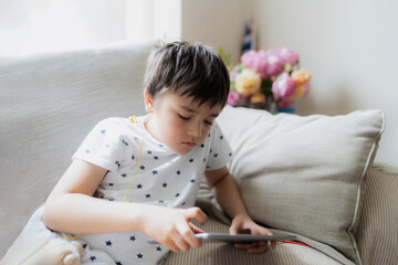 Wall Mural - High key portrait kid playing game on tablet sitting on sofa with light shining from window, Young boy  playing games on internet, Cinematic portrait Child doing homework online at home