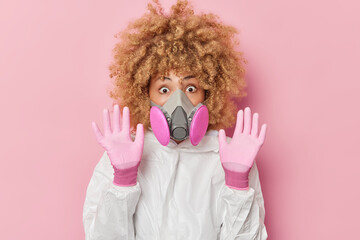 Scared impressed young woman keeps palms raised towards camera tries to protect herself from danger dressed in protective white chemical suit gloves and gas mask isolated over pink background