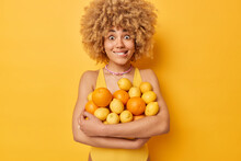 Surprised Cheerful Woman Bites Lips Carries Big Heap Of Fresh Oranges And Lemons Eats Fruits Full Of Vitamins Dressed In Casual T Shirt Isolated Over Vivid Yellow Background. People Summer Time