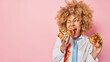 © wayhome.studio - Unhealthy junk food and cheat meal concept. Curly haired woman eats delicious hot dog and hamburger has good appetite keeps mouth widely opened dressed formally isolatd over pink wall empty space