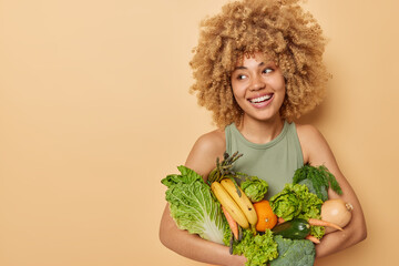 Wall Mural - Horizontal shot of beautiful woman carries variety of fresh vegetables and fruits consumes grocery full of vitamins looks away stands against beige background blank space for your promotional content