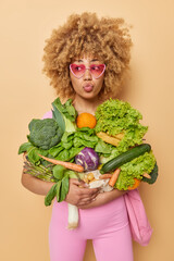 Wall Mural - Lovely curly haired young woman wears pink sunglasses carries fresh vegetables picked from garden enjoys farming poses against brown background. Homegrown bio produce and agriculture concept