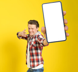 Happy to win handsome man pointing at smartphone with white empty screen, wearing red plaid shirt and jeans cellphone display mock up isolated on yellow background. Mobile app advertisement