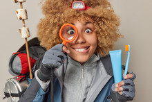 Solo Expedition Or Camping Trip. Positive Curious Curly Female Hiker Uses Magnifying Glass To Explore Something Holds Tube Of Toothpaste And Toothbrush Carries Necessary Equipment In Backpack