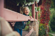 Little cute girl with blond hair laughs. Dressed in a denim suit, she stands at the wooden railing on the veranda of the cottage, decorated with flowers and vegetation.