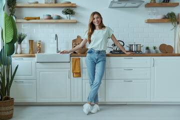 Full length of beautiful young woman leaning at the kitchen desk and smiling