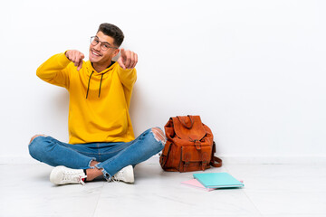 Young student caucasian man sitting one the floor isolated on white background points finger at you while smiling