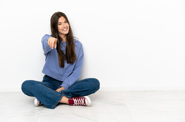 Wall Mural - Young woman sitting on the floor points finger at you with a confident expression