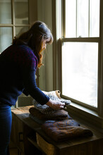 Woman Folding Hand Knit Sweaters On A Wooden Dresser By Window Light