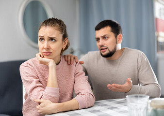 Apologetic husband calming his upset young wife while sitting at table in living room, asking for forgiveness after spat..