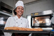 Portrait of African American female chef in white cooking uniform looking at camera with cheerful smile and proud with tray of baguette in kitchen, pastry foods professional, fresh bakery occupation.