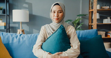 Attractive cheerful young Asian muslim beauty woman in hijab with casual sitting on couch smile looking at camera in cozy living room at home. Girl Islam social distance quarantine lifestyle concept.