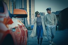 A Fashionable Young Couple With Sunglasses And Walking Beside An Orange Luxury Sports Car