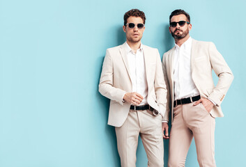 Portrait of two handsome confident stylish hipster lambersexual models. Sexy modern men dressed in white elegant suit. Fashion male posing in studio near blue wall in sunglasses